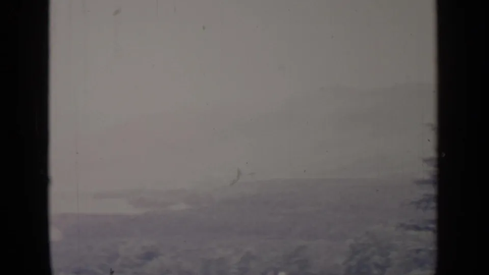 1964 8mm Greenwood Lake winter view with dark foreground framing, muted snowy hills, and a blurred evergreen edge.