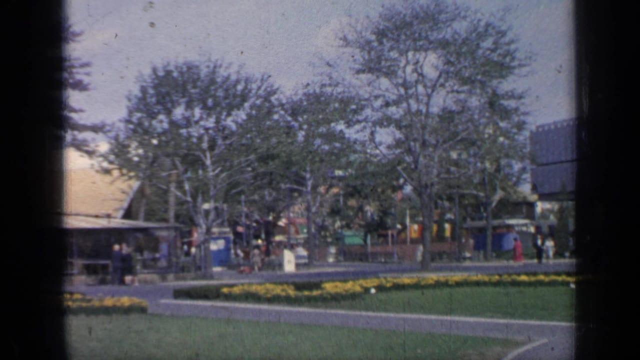 Fairground lawn and flowerbeds with trees, visitors, pavilion structures, and a restaurant area in the background.
