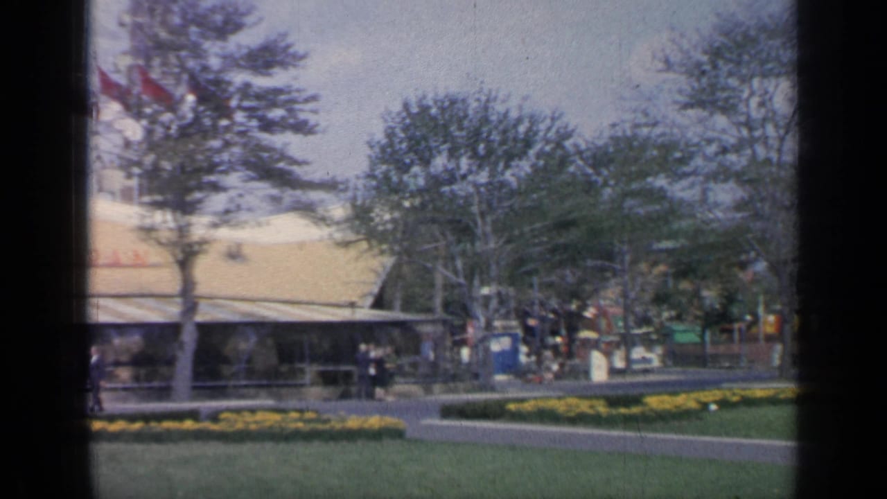 Landscaped fairground with trees, yellow flowerbeds, visitors, red flags, and pavilion buildings.