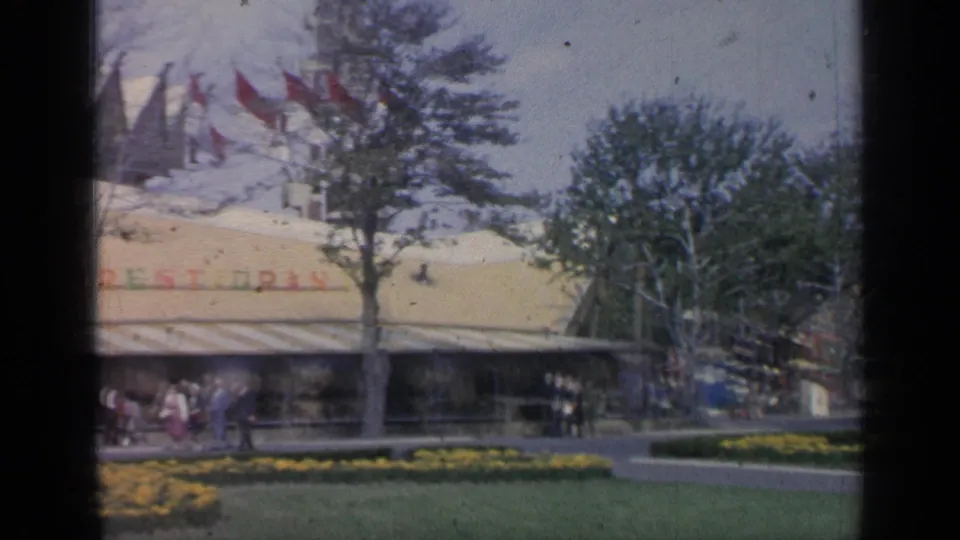 The Kynesia Restaurant pavilion appears behind trees and yellow flowerbeds, with red flags overhead and visitors gathered along the walkway.