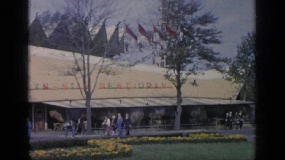 1964 Flushing Meadows view of the Kynesia Restaurant frontage, with red flags, trees, strolling visitors, and yellow flowerbeds in the foreground.