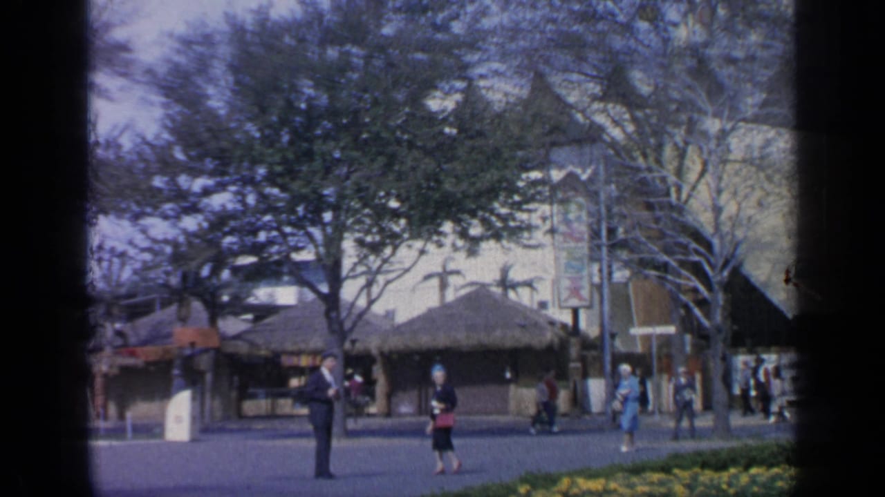 Visitors walk near thatched-roof pavilions and trees at a fairground walkway.