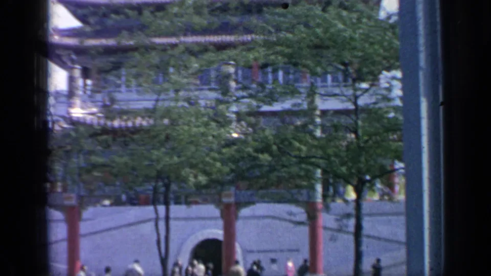 Flushing Meadows World's Fair visitors near a Chinese-style gate, with trees, red columns, and a vertical pole at frame edge.