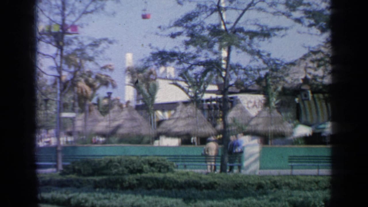 Thatched-roof fairground structures behind green fencing, trees, benches, and distant visitors.