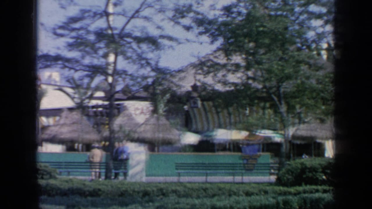 Flushing Meadows fairground scene with thatched pavilion roofs, shaded umbrellas, benches, fencing, trees, and visitors in the distance.