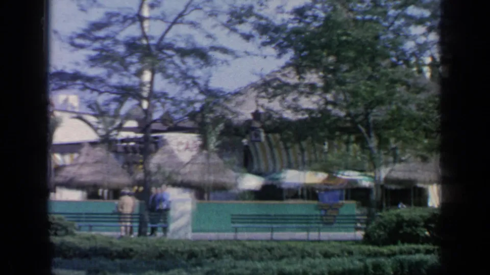 Flushing Meadows fairground scene with thatched pavilion roofs, shaded umbrellas, benches, fencing, trees, and visitors in the distance.