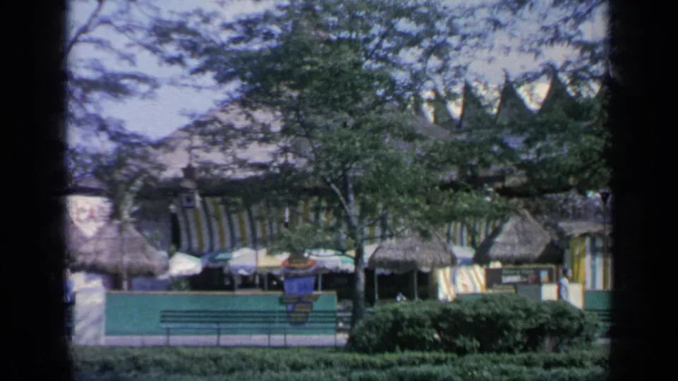 1964 World's Fair pavilion view with thatched roofing, striped awnings, trees, shrubs, benches, and green foreground fencing.