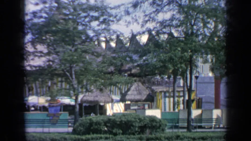 Flushing Meadows fairground view with a cable car overhead, thatched structures, umbrellas, trees, shrubs, green fencing, and benches.
