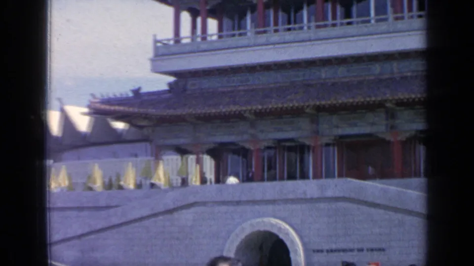 Handheld 8mm frame of the Flushing Meadows pavilion, with ornate roofline, umbrellas, terrace wall, and arched opening visible.