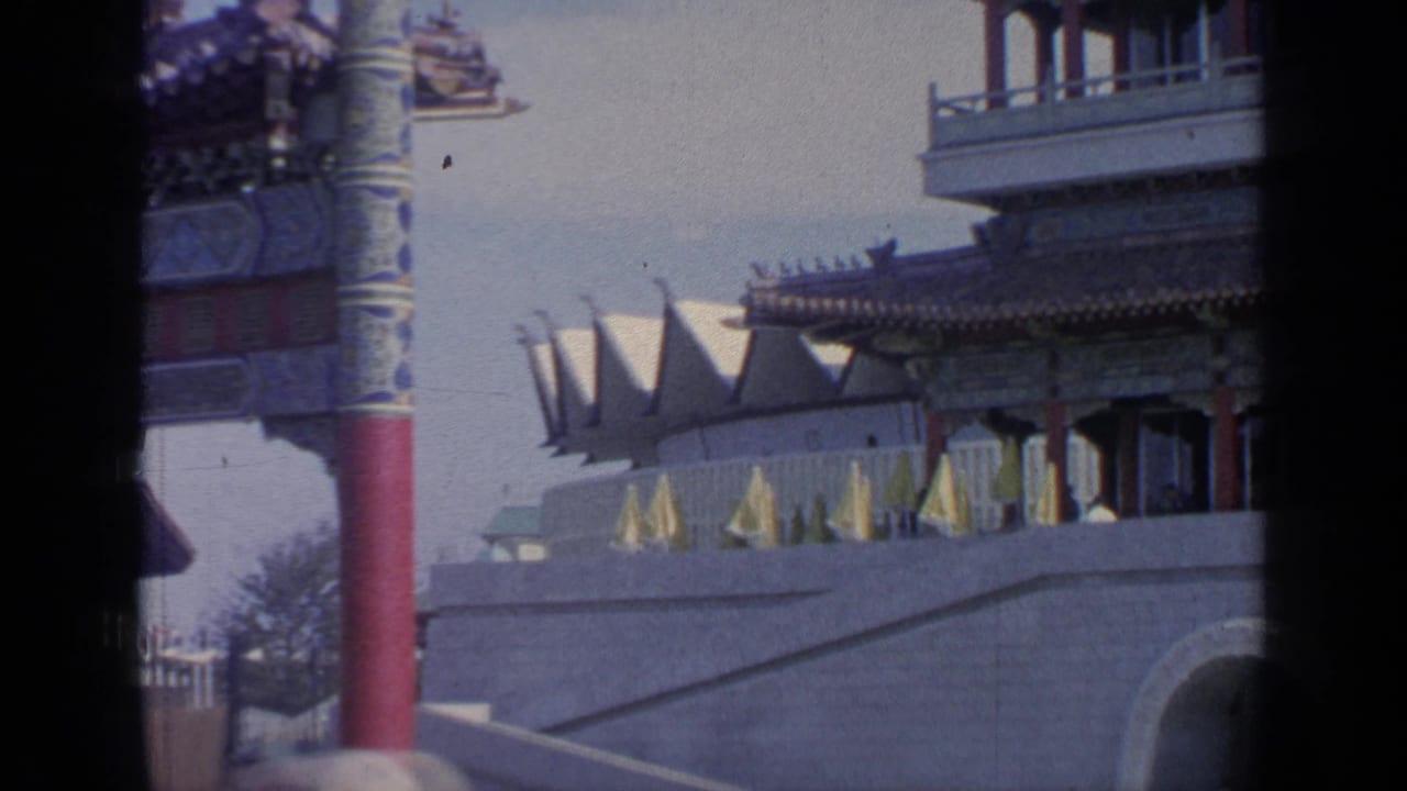 1964 Flushing Meadows World's Fair view of the Mexico Pavilion area with flags, a red column, and architectural details in a handheld 8mm frame.