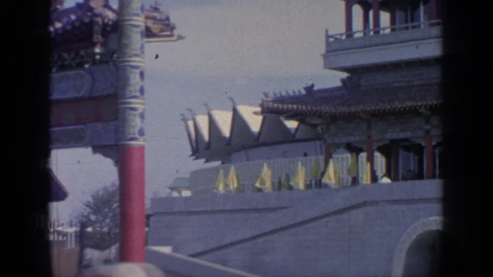 1964 Flushing Meadows World's Fair view of the Mexico Pavilion area with flags, a red column, and architectural details in a handheld 8mm frame.