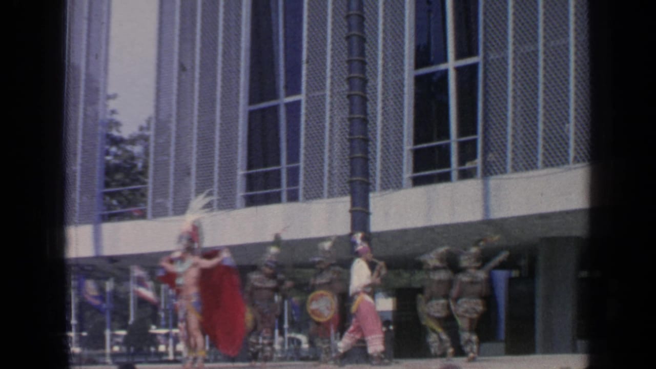 Costumed dancers with feathered headdresses, capes, and shields perform in front of a pavilion building.
