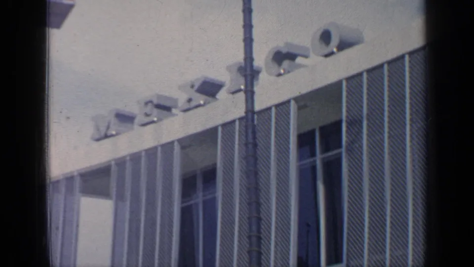 The camera frames the Mexico Pavilion facade at the 1964 New York World's Fair, with partial lettering and tall vertical windows visible.
