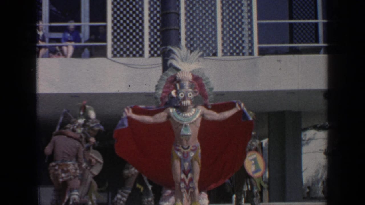 Masked dancer with feathered headdress and red cape performs before a fair building.