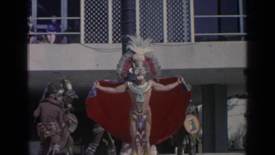 At the 1964 New York World's Fair, a masked dancer in a feathered headdress and red cape performs with spectators visible on a balcony.