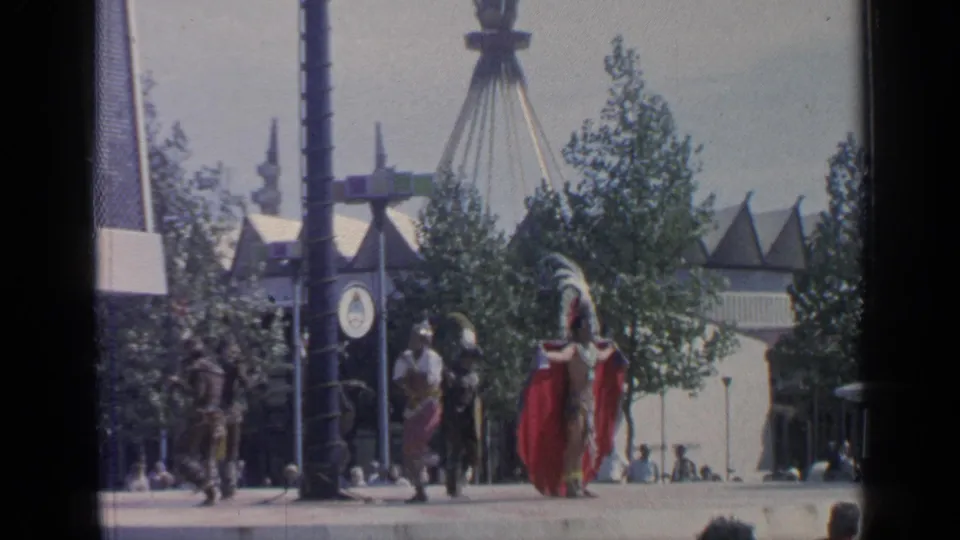 Wide 8mm view at Flushing Meadows shows costumed dancers on a fairground walkway with trees, posts, and pavilion-like roofs in the background.