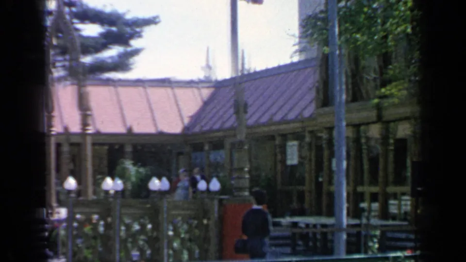 Side view near the Thailand Pavilion at the 1964 World's Fair, showing a railing, roofline, trees, lamps, and a visitor in the foreground.