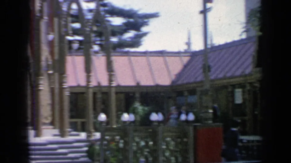 1964 Flushing Meadows home-movie frame showing the Thailand Pavilion exterior, steps, globe lamps, trees, and fair visitors.