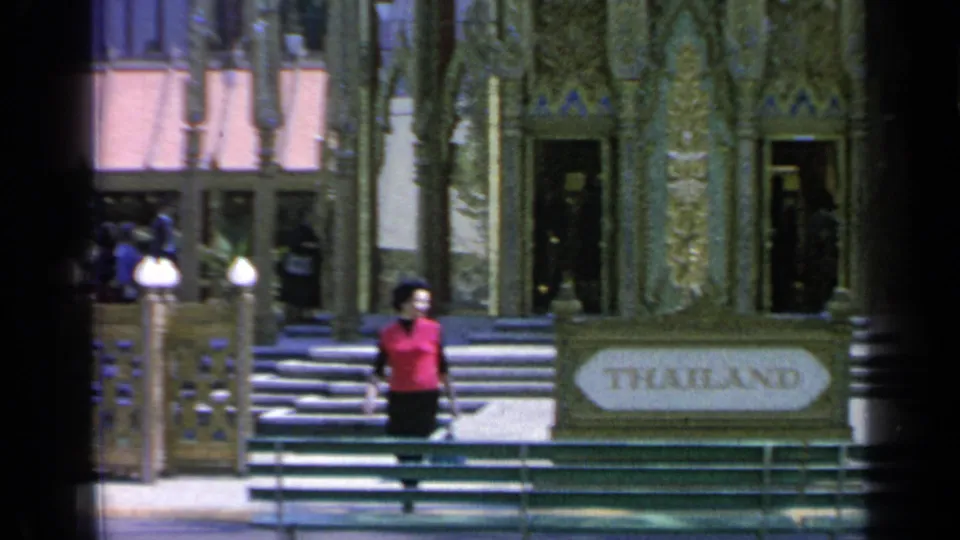 1964 Flushing Meadows frame showing the Thailand sign, pavilion steps, visitors, globe lamps, and a woman walking in front.
