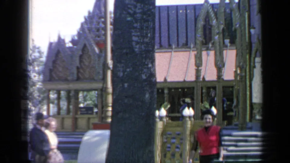 1964 World's Fair home-movie frame with a woman in red walking near the Thailand Pavilion steps and visitors nearby.