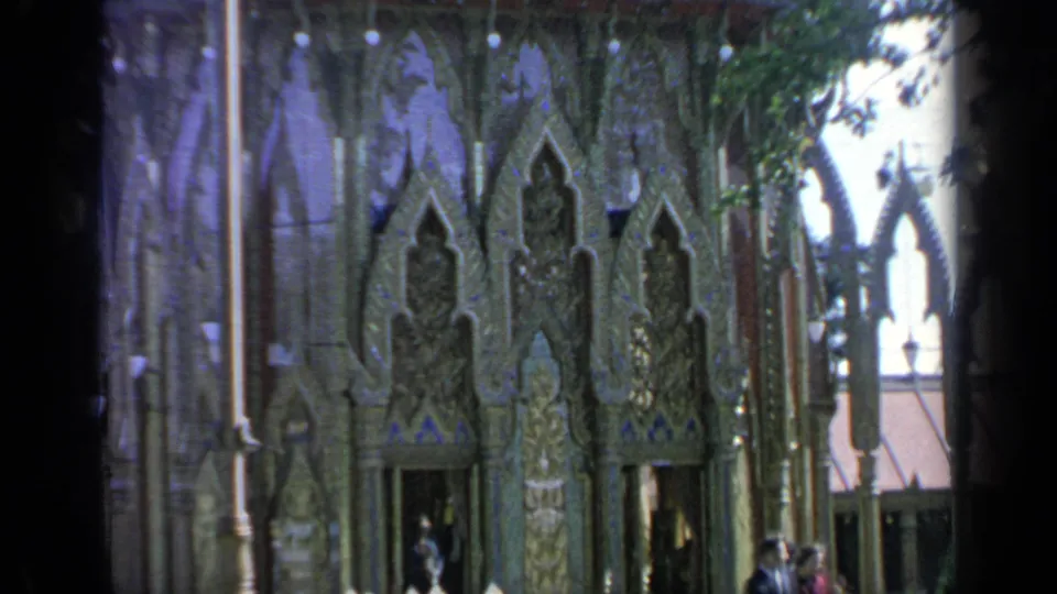 1964 Flushing Meadows frame showing the pavilion facade, decorative pointed panels, trees, and a few visitors near the base.