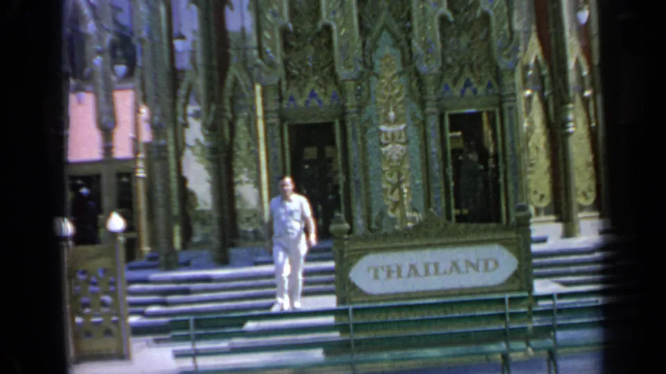 1964 Flushing Meadows home-movie frame showing a man walking by the ornate Thailand Pavilion sign and steps.