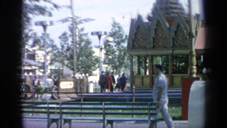 1964 World's Fair frame showing the Thailand Pavilion from the side, with visitors on the walkway and fairground lamps nearby.