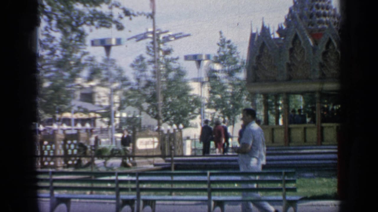 Man in a light gray suit walking past green benches and a pavilion in a sunny fairground park.