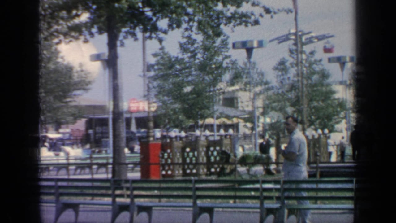 Suited man walking beside green benches with trees, lampposts and fair structures behind him.