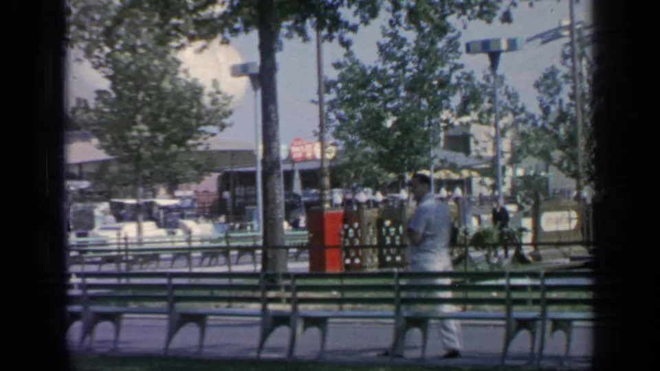 A rear view of the suited man at the 1964 Flushing Meadows fairground shows benches, trees, fencing, lampposts and distant signage.