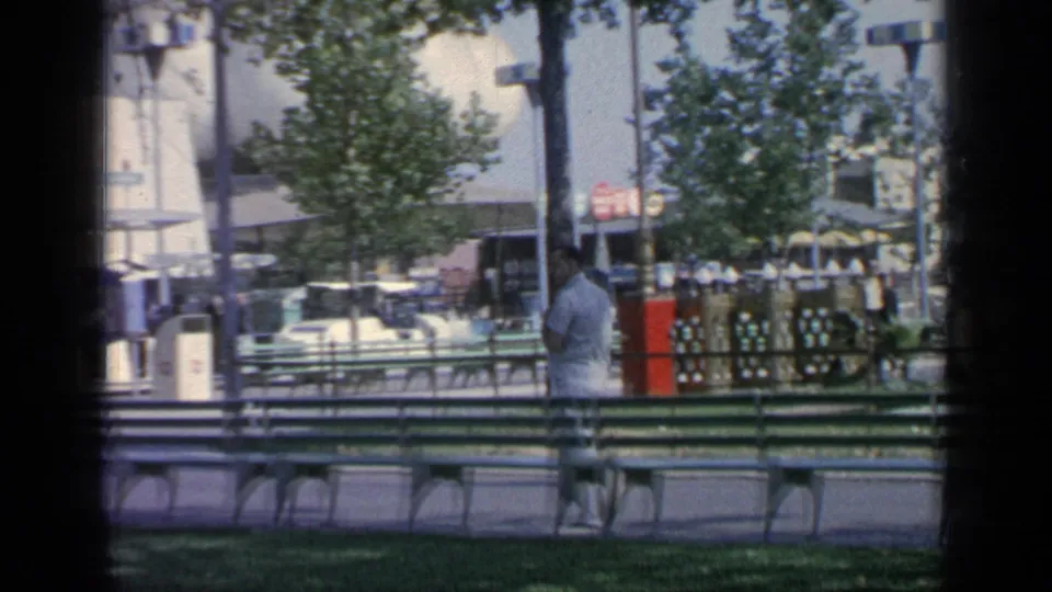 The suited man continues through the 1964 Flushing Meadows fairground, with green benches, trees, a red post and distant fair structures visible.