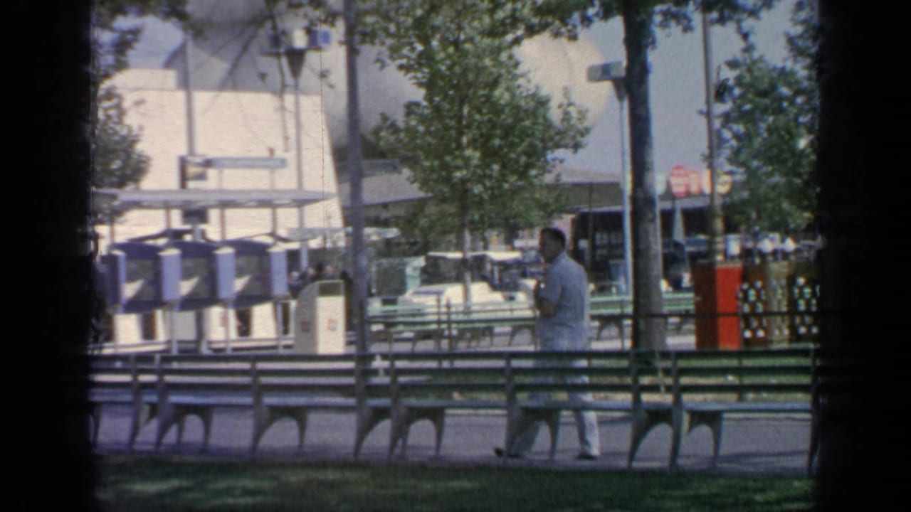 Man in a light shirt walks past green benches with trees and fair structures behind him.