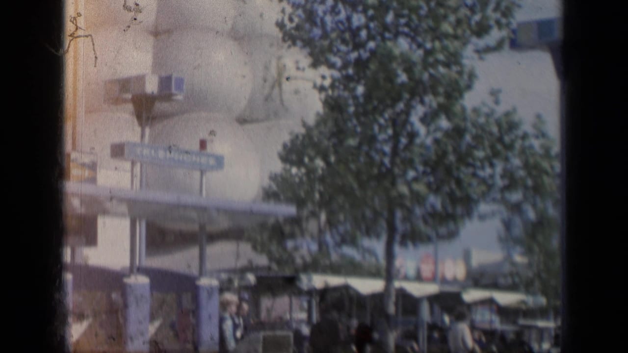 1964 Flushing Meadows fairground view with a sign, trees, visitors, and a mid-century building partly obscured by the film frame.
