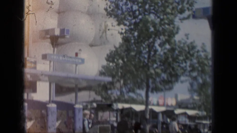 1964 Flushing Meadows fairground view with a sign, trees, visitors, and a mid-century building partly obscured by the film frame.