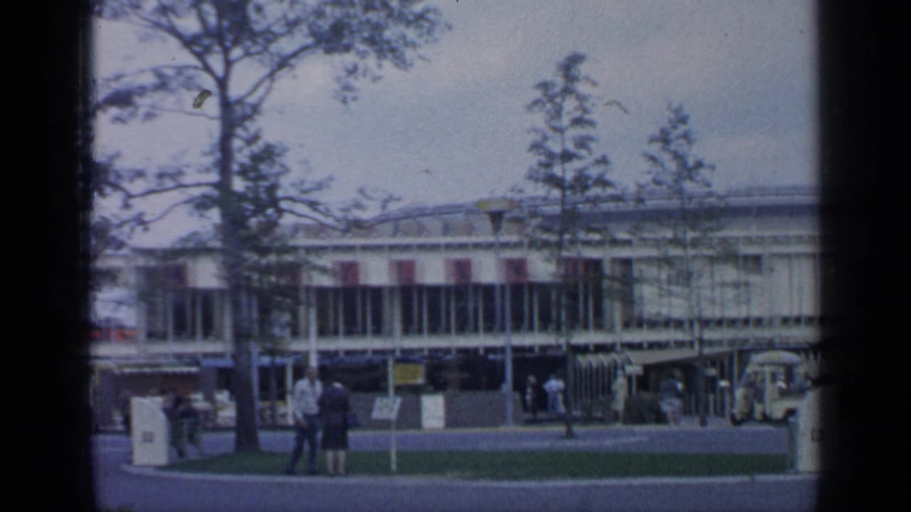 1964 World's Fair grounds at Flushing Meadows with a low modern building, trees, lawn, and visitors walking or standing nearby.