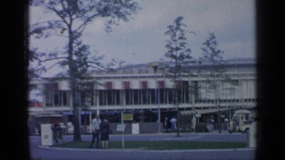 1964 World's Fair grounds at Flushing Meadows with a low modern building, trees, lawn, and visitors walking or standing nearby.