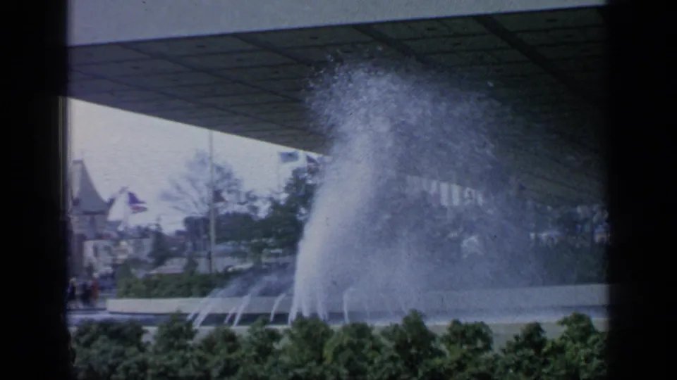 Large fountain at the 1964 Flushing Meadows fairgrounds, with water spray, plantings, visitors, and distant pavilion-like structures.