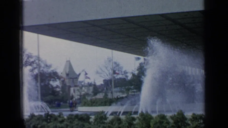 1964 Flushing Meadows fountain view with water jets, landscaped plantings, pedestrians, and distant fair architecture under an 8mm frame shadow.