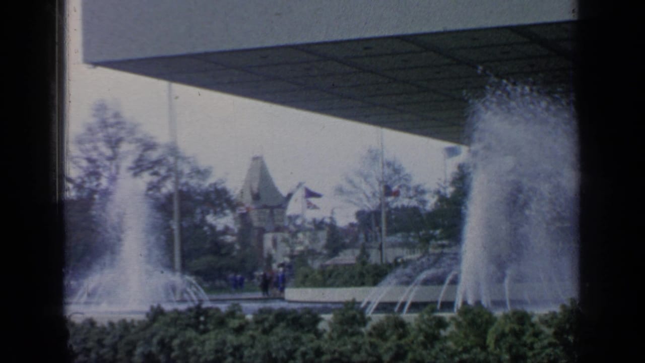Fountains spray near a modern pavilion roof with fairground buildings and visitors in the background.