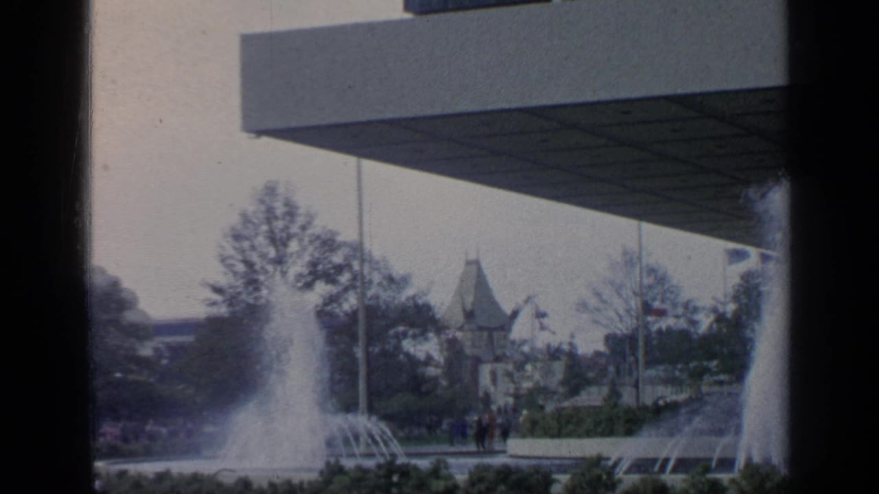 Fountain jets rise beside trees and distant fairground buildings under a large overhang.