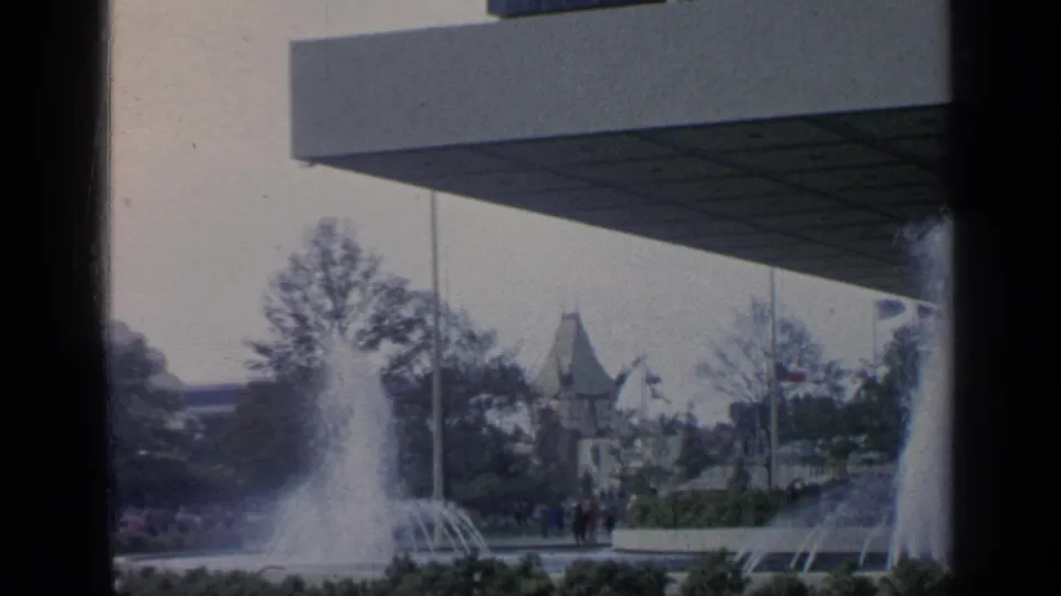 Visitor-shot 1964 World's Fair scene with fountains, a broad pavilion roof, trees, distant structures, and small groups of people.