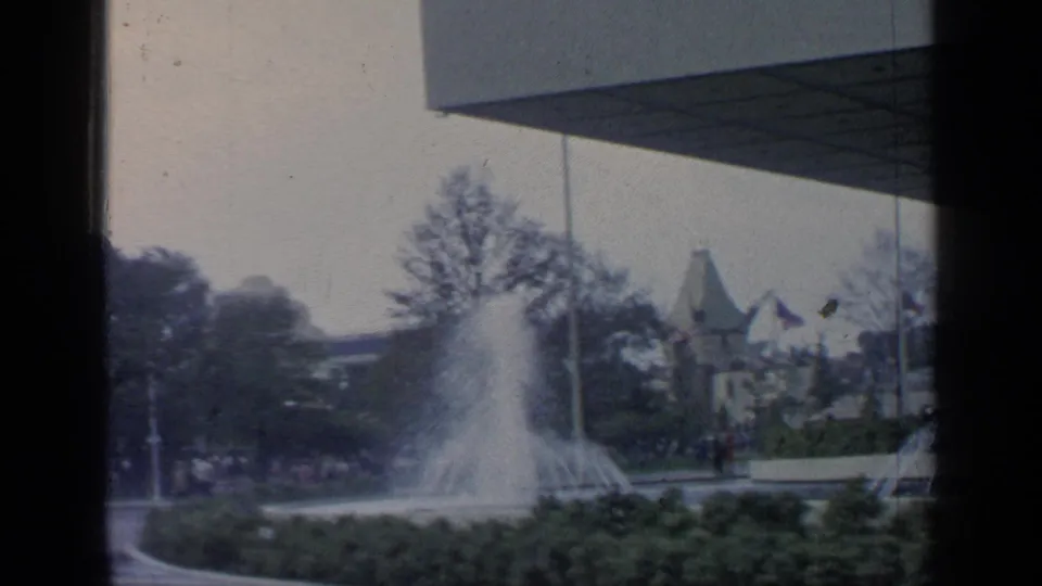 1960s Flushing Meadows view of fountain spray, low plantings, trees, distant fair structures, and a modern roofline.