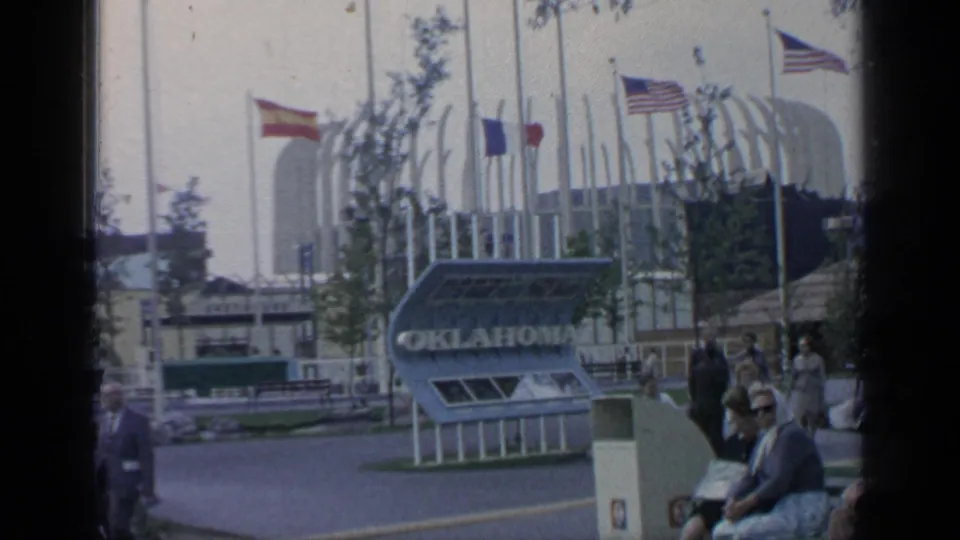 1964 World's Fair frame showing the Oklahoma pavilion sign, flags, visitors, a walkway, and curved fairground architecture.
