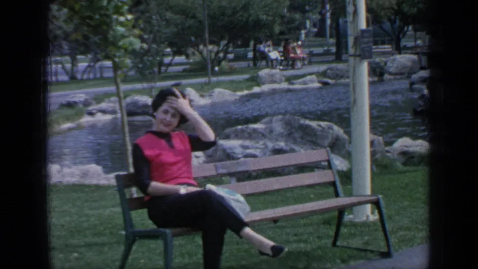 Woman in a red sleeveless top sits on a park bench near rocks, grass, trees, and a pond at the fairgrounds.