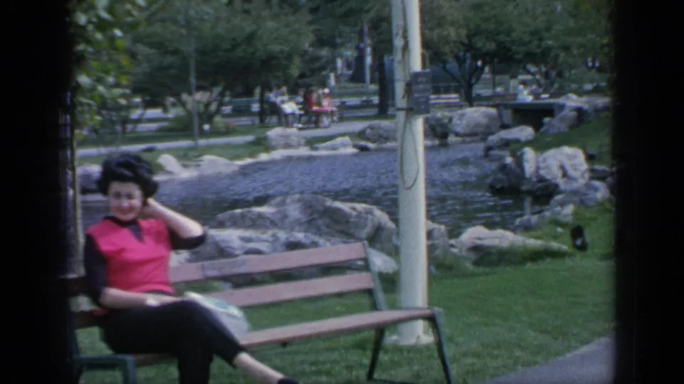 Bench portrait of a woman in a red top beside a landscaped pond at Flushing Meadows, with trees and paths behind her.