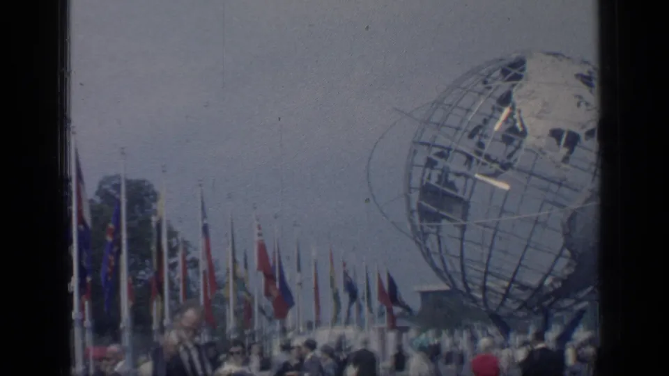 Visitor-shot 8mm frame shows part of the Unisphere, rows of flags, and bundled fairgoers at Flushing Meadows.