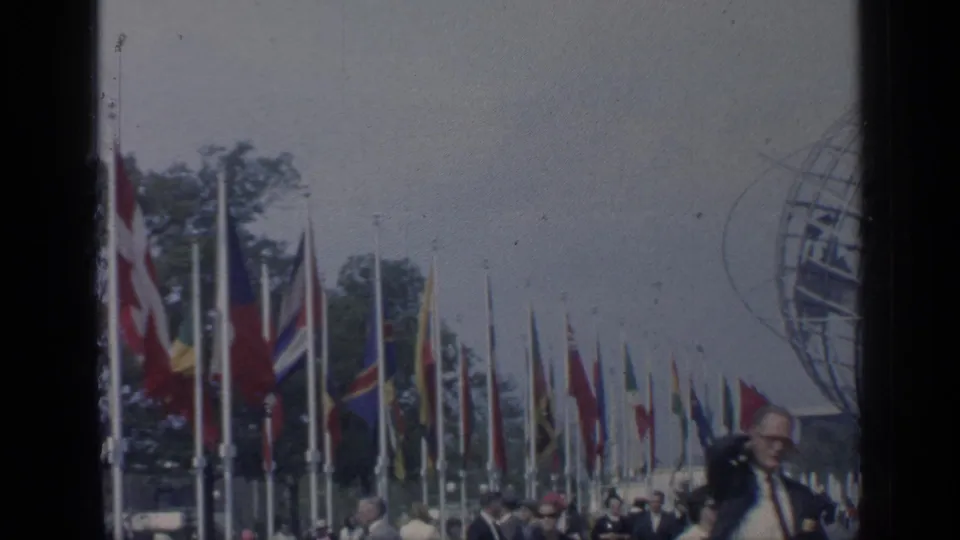 Rows of flags and public visitors stand in front of a partly cropped Unisphere at Flushing Meadows in 1964 footage.