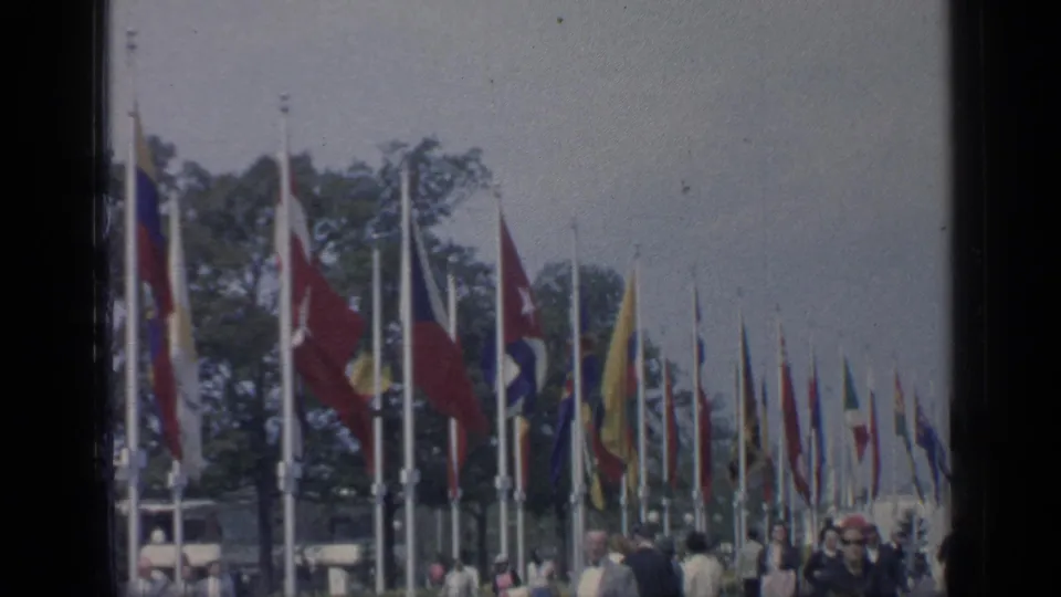 1964 Flushing Meadows fairground view with rows of national flags, trees, and visitors moving through the walkway.
