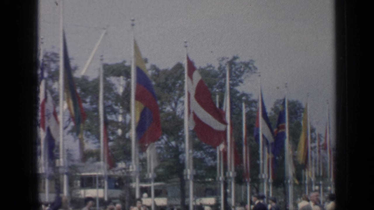 National flags flutter above a crowd near trees and fair structures.