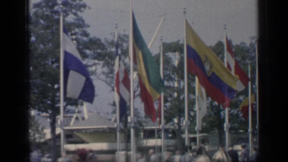 Closer 8mm view of multiple national flags on tall poles with a fairground crowd below in Flushing Meadows.